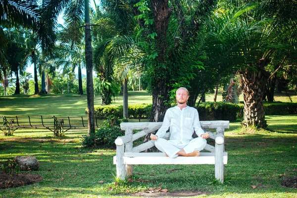 Man meditating on bench in park. Young male in white clothing me