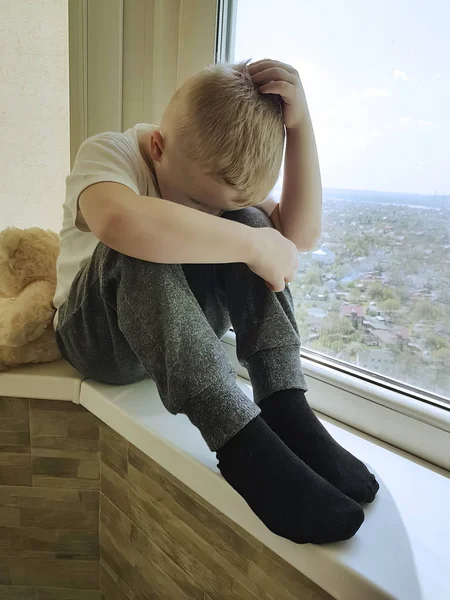 little sad boy sits on a window with a teddy bear
