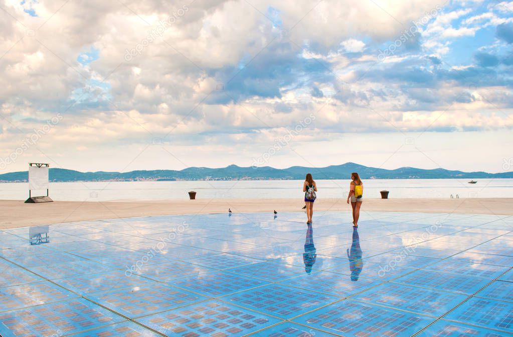 Two girls walking on top of Sun Salutation installation made of solar photovoltaic cells near the sea shore against the background of a hill range and dramatic cloudy morning sky. Zadar, Croatia