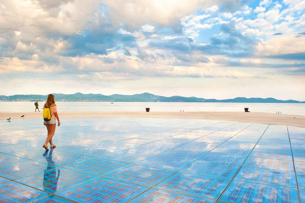 A girl walking on top of Sun Salutation installation made of solar photovoltaic cells near the sea shore against the background of a hill range and dramatic cloudy morning sky. Zadar, Croatia