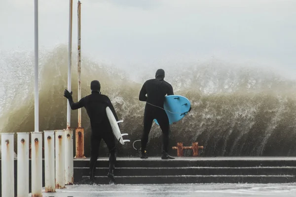 Två surfare i våtdräkt med surfbräda på en stadskaj mitt i kraftfulla vågor i Australien. Begreppet: konfrontation mellan elementen och människan, extrema sporter och hobbyer, natur vs människa. Bakifrån — Stockfoto