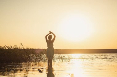 Gölün kıyısında uzun beyaz bir elbiseyle gün batımını izleyen bir kadın. Arkadan çekilmiş, yüzü olmayan bir fotoğraf. Seçici odak.
