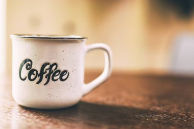 Enamel beyaz metal kupası. Üzerinde yazılı kahve yazılı. Arka planda bulanık tahta masa. Siyah arka planda beyaz bir fincan kahve. Metin veya reklam için yer