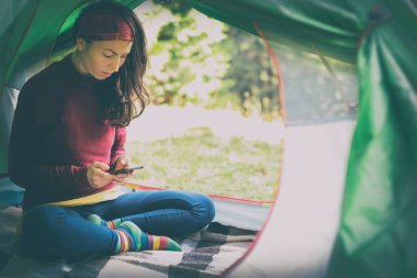 Ormana bakan bir çadırdaki Avrupalı kadın elinde bir telefon tutuyor. Tatilde ve tatilde her zaman temasta olmak için cihazları ve interneti olan modern bir bayram..