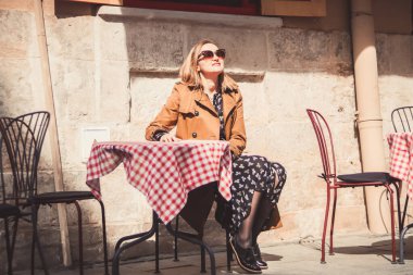 Açık hava kafeteryasında oturan ve güneşli bahar günlerini seven kahverengi paltolu güzel sarışın kadın düşünceli görünüyor..
