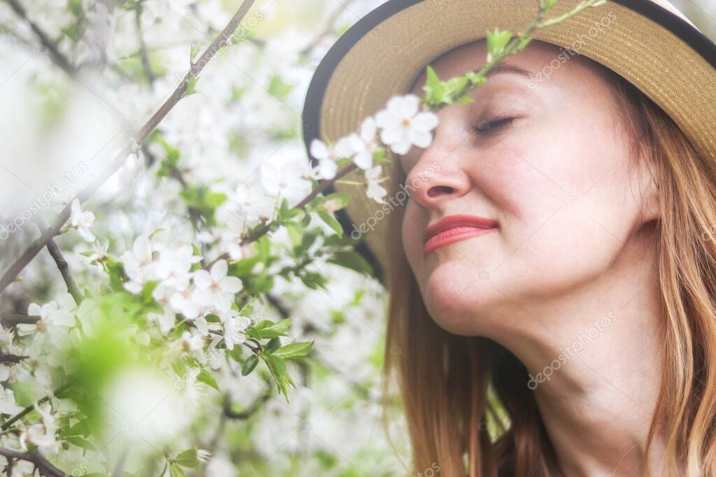 Beautiful  girl in straw hat sniffs blossom branch.  Portrait of a beautiful young blonde woman with long hair in a blue dress on background of white cherry blossoms in spring. Spring season concept.