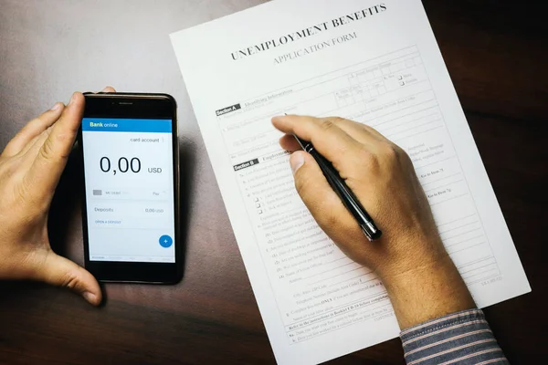 A man checks her account balance on phone. Lack of money, filling out unemployment benefits form.  All screen graphics are made up. Concept of coronavirus and stay at home order impact on economy