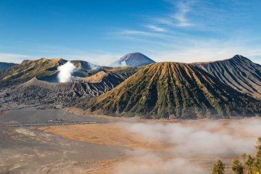 Bromo Dağı volkanı, Doğu Java adası, Endonezya. Bulutlar vadiyi kaplıyor, koninin eteğindeki Luhur Poten Tapınağı. Arka planda mavi gökyüzüne karşı yükselen başka bir volkanın gazları..