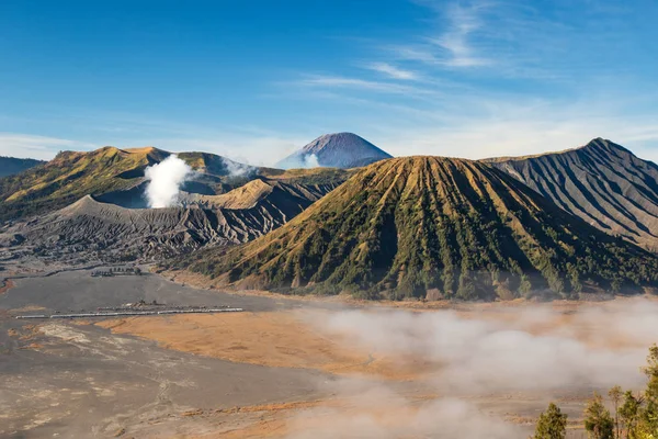 Bromo Dağı volkanı, Doğu Java adası, Endonezya. Bulutlar vadiyi kaplıyor, koninin eteğindeki Luhur Poten Tapınağı. Arka planda mavi gökyüzüne karşı yükselen başka bir volkanın gazları..