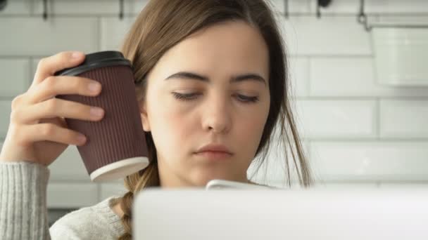 A menina bebe café, escreve sms em um smartphone e trabalha para um laptop. Menina atraente trabalhando — Vídeo de Stock