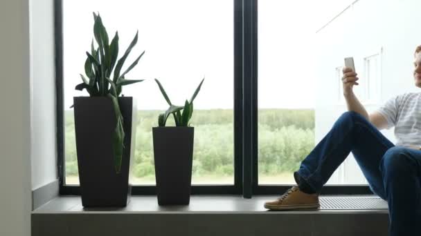 El hombre escribe un SMS y mira por la ventana del aeropuerto. El tipo está hablando en mensajeros. Un joven en un smartphone juega. Un tipo escribe un mensaje de texto a una chica y sueña. desplazamiento imágenes phon — Vídeo de stock