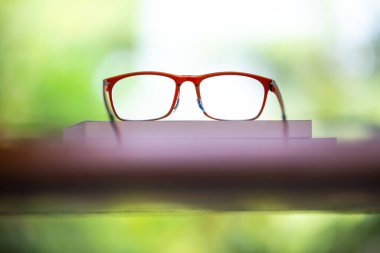 Orange eyeglasses with books on wooden table, Bokeh garden background, Close up & Macro shot, Selective focus, Stationery concept