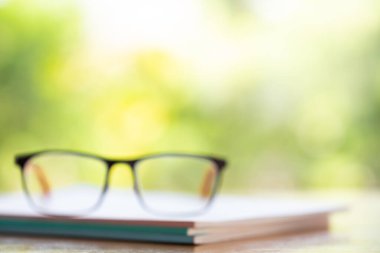 Blurred black eyeglasses with three white notebooks on wooden table, Bokeh garden background, Close up & Macro shot, Stationery concept