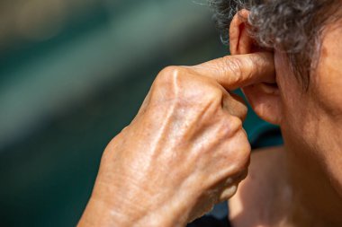 Senior woman plugging index finger into her right ear, Grey curly hairs, Swimming pool background, Close up & Macro shot, Selective focus, Asian Body part, Healthcare concept, Symptom of hearing loss