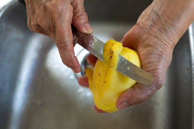 Yaşlı kadının sağ eli bıçak kullanıyor lavabonun arka planında tatlı sarı mango soyuyor yakın çekim, seçici odak, Tayland geleneksel meyvesi