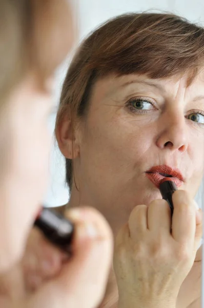 Head portrait in the mirror of a middle-aged brunette woman in front of the mirror with lipstick put on her lips.