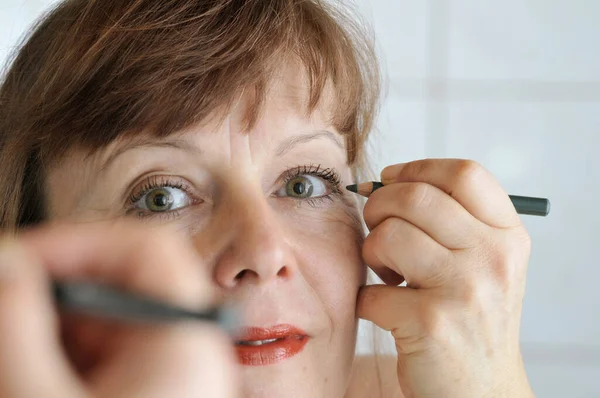 Head portrait in the mirror of a brunette middle-aged woman pulling an eyeliner with a pencil in front of the mirror.