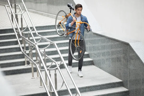 Un tipo con chaqueta de mezclilla bajando las escaleras sosteniendo una bicicleta naranja. Estudiante con la bicicleta — Foto de Stock