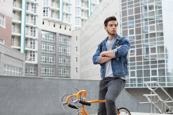 El tipo va a la ciudad en bicicleta con chaqueta vaquera azul. joven un naranja fijar bicicleta — Foto de Stock