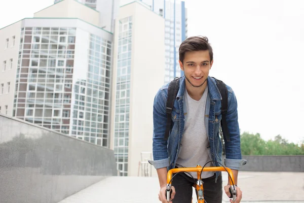 El tipo va a la ciudad en bicicleta con chaqueta vaquera azul. joven un naranja fijar bicicleta — Foto de Stock