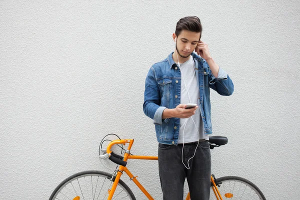 El tipo con una chaqueta de mezclilla azul de pie sobre el fondo de la pared. Un joven cerca de la bicicleta naranja. Estudiante sonriente con bolsa mirando el teléfono — Foto de Stock