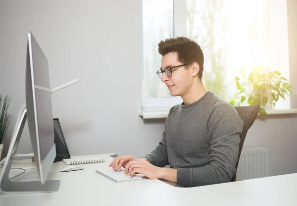 Un joven trabajador sentado en una oficina en la computadora. Freelancer con camisa blanca y gafas. El diseñador se sienta frente a la ventana en el lugar de trabajo . — Foto de Stock