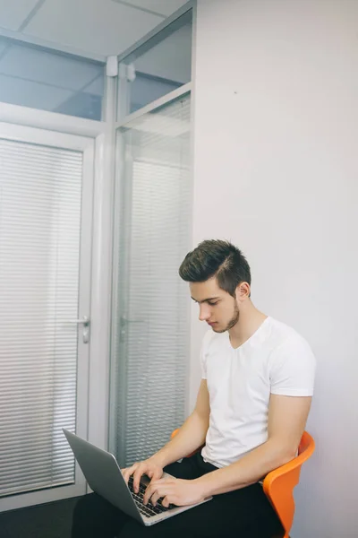 Νέος εργαζόμενος που κάθεται σε ένα γραφείο στον υπολογιστή. Ελεύθερος επαγγελματίας σε ένα λευκό πουκάμισο. Ο σχεδιαστής κάθεται στον εργασιακό χώρο. Φοιτητής με φορητό υπολογιστή. Πορτοκαλί καρέκλα — Φωτογραφία Αρχείου