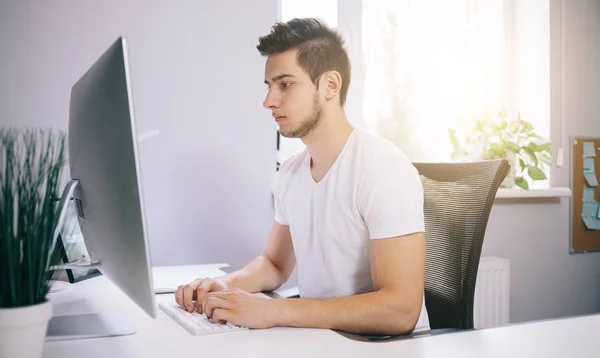 Молодой работник сидит в офисе за компьютером. Фрилансер в белой рубашке. Дизайнер сидит перед окном на рабочем месте . — стоковое фото