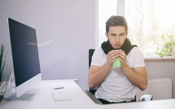 Un joven trabajador resfriado. Freelancer en bufanda y taza de té en la oficina. Diseñar la temperatura en el lugar de trabajo . — Foto de Stock