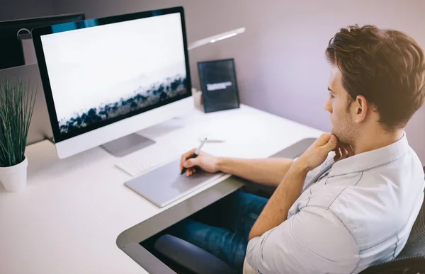Junger Arbeiter sitzt in einem Büro am Computer. Freiberufler im blauen Hemd. der Designer sitzt am Arbeitsplatz vor dem Fenster. — Stockfoto