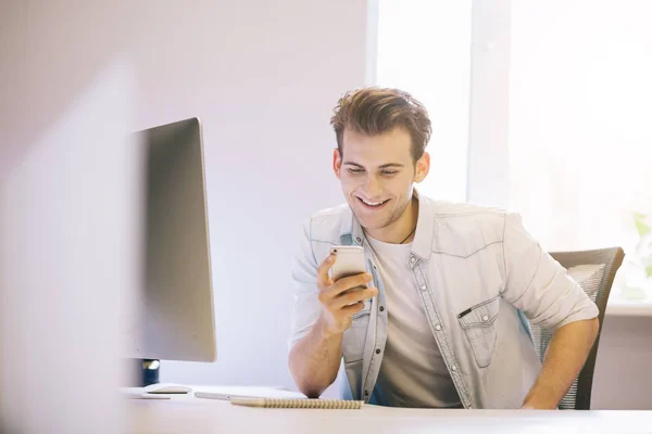 Sonriente hombre hablando en el teléfono móvil mientras se utiliza el ordenador portátil en el escritorio en el estudio . — Foto de Stock