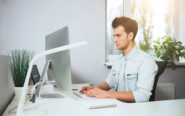 Un joven trabajador sentado en una oficina en la computadora. Freelancer con camisa blanca. El diseñador se sienta frente a la ventana en el lugar de trabajo . — Foto de Stock