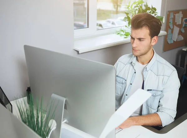 Νέος εργαζόμενος που κάθεται σε ένα γραφείο στον υπολογιστή. Ελεύθερος επαγγελματίας σε ένα λευκό πουκάμισο. Ο σχεδιαστής που κάθεται μπροστά από το παράθυρο στο χώρο εργασίας. — Φωτογραφία Αρχείου