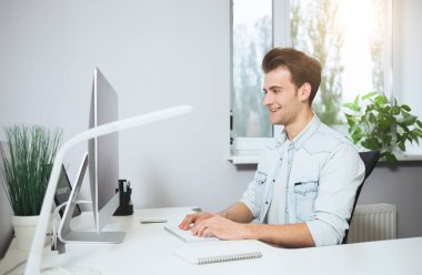 Bir ofiste bilgisayar başında oturan genç bir işçi. Freelancer Beyaz gömlekli. Tasarımcı işyerinde pencerenin önünde oturur.