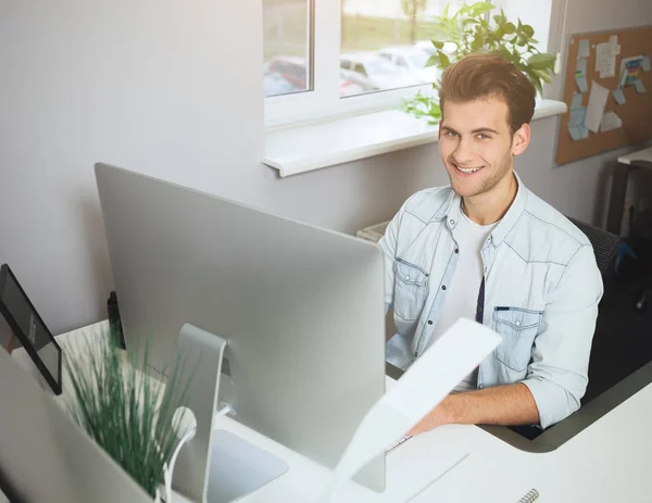 Young worker sitting in an office at the computer. Freelancer in a white shirt. The designer sits in front of window in the workplace.