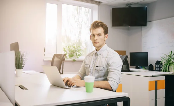 Молодой парень сидит за компьютером в офисе с окнами за спиной — стоковое фото