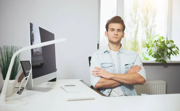 Un joven trabajador sentado en una oficina en la computadora. Freelancer con camisa blanca. El diseñador se sienta frente a la ventana en el lugar de trabajo . — Foto de Stock