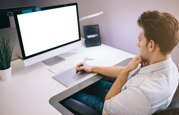 Νέος εργαζόμενος που κάθεται σε ένα γραφείο στον υπολογιστή. Ελεύθερος επαγγελματίας σε ένα μπλε πουκάμισο. Ο σχεδιαστής που κάθεται μπροστά από το παράθυρο στο χώρο εργασίας. — Φωτογραφία Αρχείου