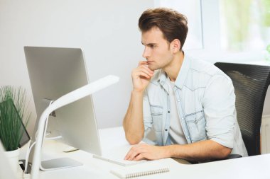 Genç bir programcı kameraya bakıyor. Grafik tasarımcı gelecekteki projeler hakkında düşünüyor. Genç adam ofis bilgisayara windows ile arkasında oturuyor