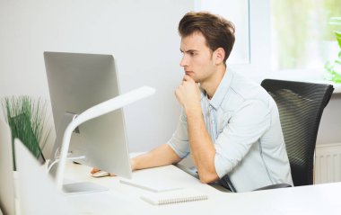 Genç bir programcı kameraya bakıyor. Grafik tasarımcı gelecekteki projeler hakkında düşünüyor. Genç adam ofis bilgisayara windows ile arkasında oturuyor