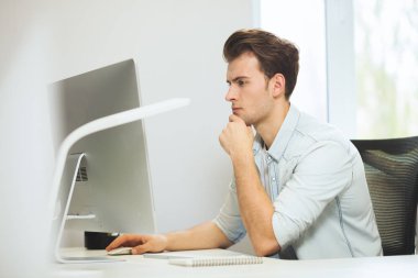 Genç bir programcı kameraya bakıyor. Grafik tasarımcı gelecekteki projeler hakkında düşünüyor. Genç adam ofis bilgisayara windows ile arkasında oturuyor