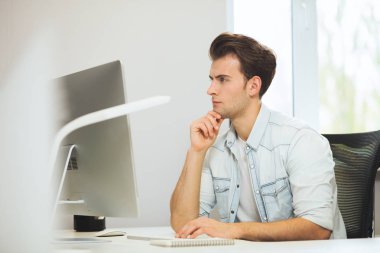 Genç bir programcı kameraya bakıyor. Grafik tasarımcı gelecekteki projeler hakkında düşünüyor. Genç adam ofis bilgisayara windows ile arkasında oturuyor