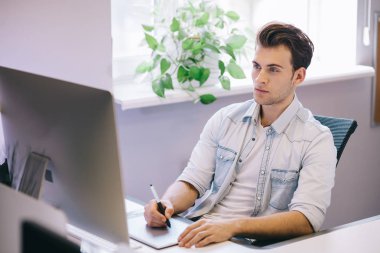Bir ofiste bilgisayar başında oturan genç bir işçi. Freelancer mavi gömlekli. Tasarımcı işyerinde pencerenin önünde oturur.