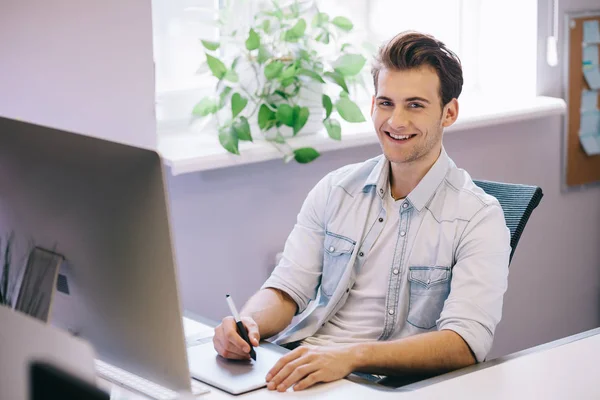 Un joven trabajador sentado en una oficina en la computadora. Freelancer con camisa azul. El diseñador se sienta frente a la ventana en el lugar de trabajo . — Foto de Stock