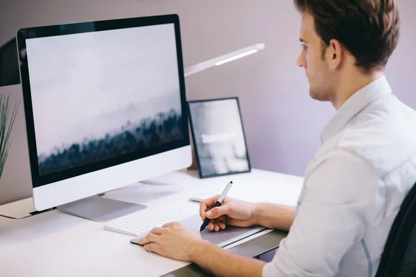 Jongeren zitten in een kantoor op de computer. Freelancer in een blauw shirt. De designer zit voor raam op de werkplek. — Stockfoto