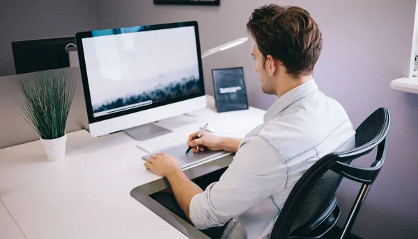 Junger Arbeiter sitzt in einem Büro am Computer. Freiberufler im blauen Hemd. der Designer sitzt am Arbeitsplatz vor dem Fenster. — Stockfoto