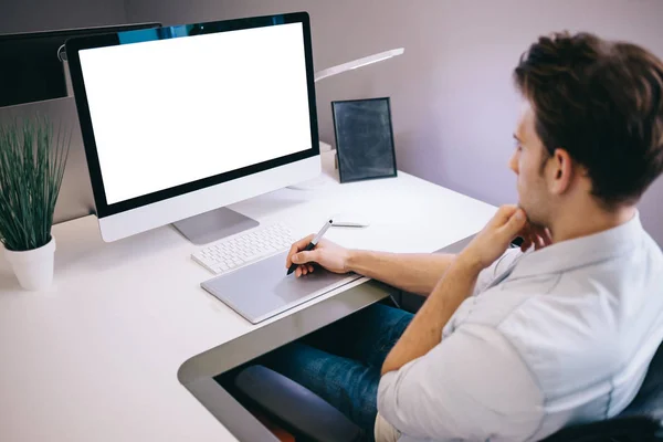 Молодой работник сидит в офисе за компьютером. Фрилансер в голубой рубашке. Дизайнер сидит перед окном на рабочем месте . — стоковое фото