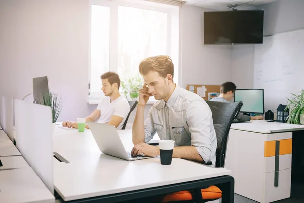 Ein junger Programmierer schaut auf den Computer. denkt die Grafikdesignerin an die Zukunftsprojekte. der junge Kerl sitzt im Büro am Computer mit den Fenstern im Rücken — Stockfoto