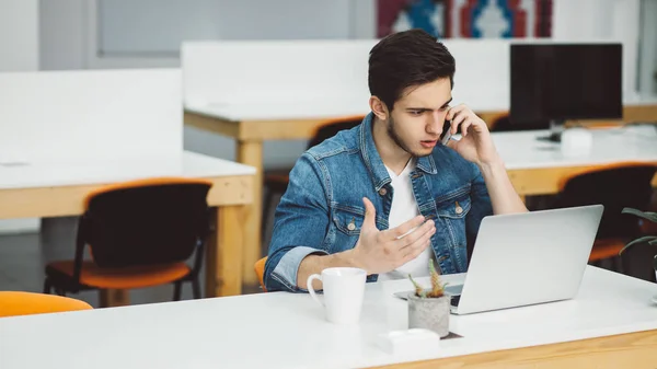 Joven hombre ocupado con barba trabajando en el ordenador portátil y el uso de teléfono móvil — Foto de Stock