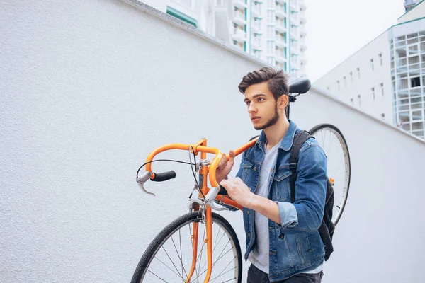 Der Typ in der blauen Jeansjacke trägt ein schulterorangefarbenes Fahrrad. ein junger Mann auf einem orangefarbenen Fahrrad — Stockfoto